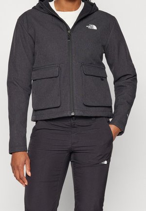 Mand iført sort North Face lynlåstrøje med klaplommer og sorte North Face bukser, hånd i lommen, hvid skjorte under.