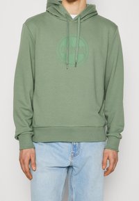 Man i en ljusgrön hoodie med en cirkulär logotyp och dragsnören, tillsammans med ljusblå jeans, stående mot en enfärgad bakgrund.