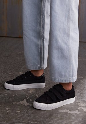 Personne portant des baskets noires avec des semelles blanches et un pantalon droit de couleur claire, debout sur un sol en béton texturé.