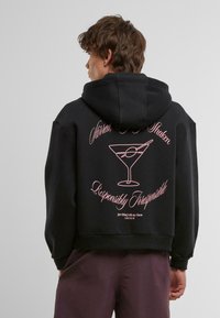 Felpa nera con cappuccio caratterizzata da testo grafico rosa e un design di un bicchiere di martini sul retro. Tessuto morbido, vestibilità rilassata, stile corto.
