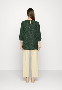 Top verde com padrão floral e mangas 3/4, detalhe de abertura nas costas, combinado com calças largas em creme e saltos bege.