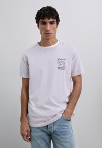 Joven con una camiseta gráfica de color púrpura claro que dice "Scalpers Lounge Bar", combinada con jeans de color azul claro, de pie frente a un fondo liso.