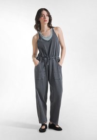 Mono de algodón gris con un corte holgado, cintura ajustable con cordón, bolsillos laterales y piernas anchas, combinado con una camiseta interior de tirantes azul claro.