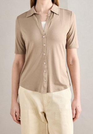 Personne portant une chemise beige à manches courtes avec des boutons par-dessus un haut blanc et un pantalon couleur crème, debout devant un fond uni.