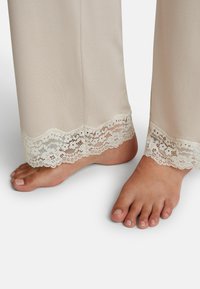 Béžové saténové pyžamo kalhoty s jemným krajkovým lemem na spodním okraji. Nohy viditelné; tón pleti je světlý, nehty nejsou nalakované.