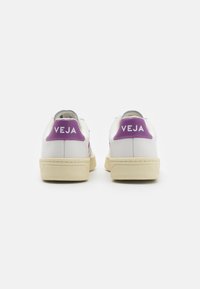 Balti sportbačiai su tekstūruotu paviršiumi, violetinė kulno juostelė su "VEJA" logotipu ir kreminės spalvos padu. Minimalistinis dizainas ir lygus paviršius.