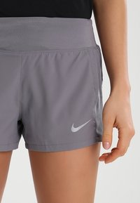 Pantalones cortos deportivos grises con una amplia cinturilla elástica, bolsillos laterales y detalles perforados. Presenta un pequeño logo plateado de Nike en la parte frontal.
