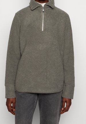 Homme portant un pull en polaire sherpa gris avec un col à fermeture éclair sur le devant et des manches longues avec poignets, associé à un jean gris foncé.