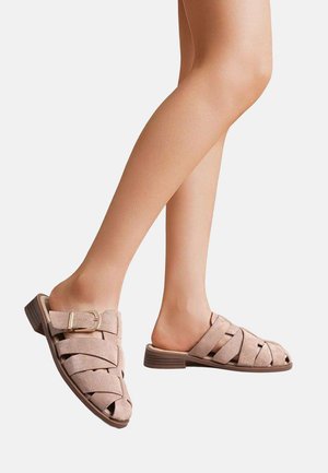 Jambes portant des sandales beige en daim tissé, à enfiler, avec des talons bas empilés et un détail de boucle sur un fond blanc.