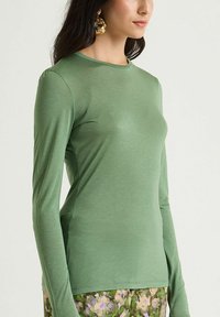 Top aderente a manica lunga in tessuto verde oliva tinta unita con texture liscia e scollo rotondo. Abbinato a pantaloni a stampa floreale.