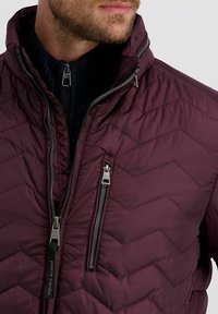 Man in een donkerpaarse gewatteerde jas met een ritszak op de borst over een zwarte geribbelde ritstrui.