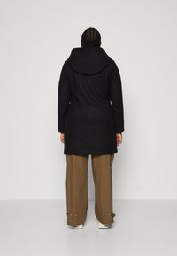 Person mit geflochtenem Haar, der einen schwarzen Kapuzenmantel, braune weite Hosen und weiße Turnschuhe trägt, mit dem Rücken zu einer schlichten Hintergrund.