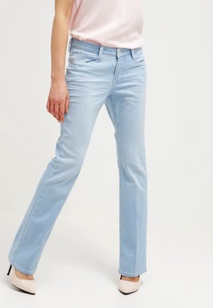 Femme portant un jean droit bleu clair et des chaussures à talons beige, debout avec une jambe légèrement pliée sur un fond uni.