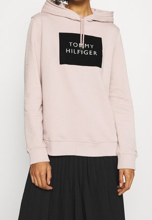 Osoba nosząca jasnoróżową bluzę z kapturem z logo "Tommy Hilfiger" z przodu oraz czarną plisowaną spódnicę, widoczna od szyi do bioder.