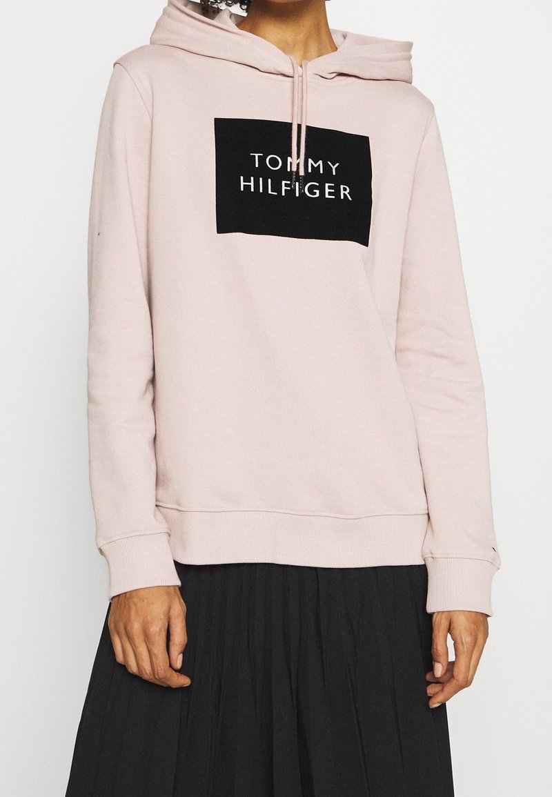 Personne portant un hoodie rose clair avec le logo "Tommy Hilfiger" sur le devant et une jupe plissée noire, montrée du cou aux hanches.