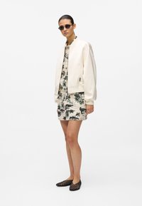 Blouson aviateur crème porté sur une robe chemise à motifs avec des palmiers, associé à des ballerines en maille noire et des lunettes de soleil rondes.