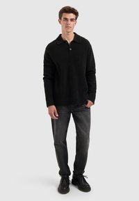 Zwart textuur pullover met een kraag en een enkele knop. Gecombineerd met grijze spijkerbroek en zwarte schoenen, staande tegen een grijze achtergrond.