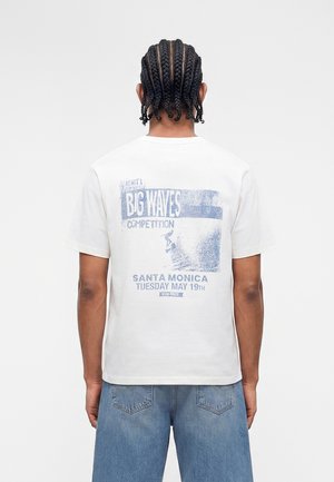 Person med flettet hår iført en hvid T-shirt med blå "Big Waves Competition Santa Monica May 19" grafik og blå jeans, set bagfra.