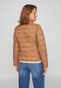 Pruun pufferi jope, millel on tikandiga disain, kõrge krae, tõmblukk ja pikad varrukad, kantud koos siniste teksadega.