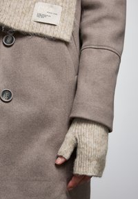 Cappotto in misto lana beige con un colletto lavorato a maglia crema, dotato di etichetta e chiusura a bottone. La manica mostra un guanto senza dita in un colore simile.