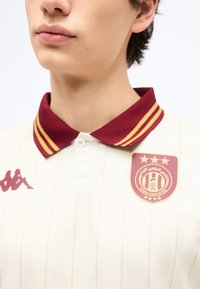 Νεαρός που φοράει αθλητική φανέλα κρεμ με μπορντό γιακά με κίτρινες ρίγες και κεντημένο έμβλημα ομάδας στο στήθος.