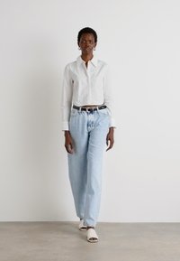 Camicia bianca con ricami, jeans a vita alta azzurri chiari e mules bianchi. Modella in piedi su uno sfondo neutro.