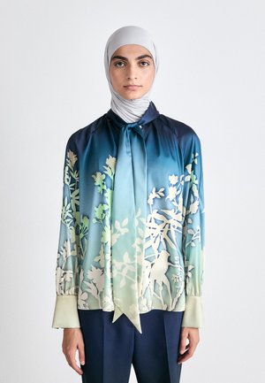 Blusa de seda con un degradado de color azul a verde, estampado floral, detalle de cuello con lazo y mangas largas; combinada con pantalones oscuros.