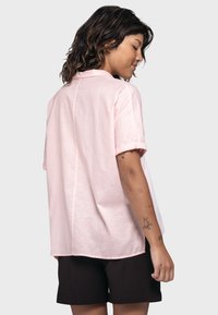 Helles rosa kurzärmeliges Hemd mit Kragen, gerader Schnitt und sichtbaren Nähten entlang des Rückens. Kombiniert mit dunklen Shorts.