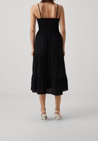 Robe midi noire avec corsage froncé et fines bretelles, présentant des volants superposés à l'ourlet. Portée avec des sandales à talons blanches.