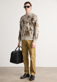 Beige és barna mintás kötött pulóver, világosbarna nadrág, fekete cipők és fekete bőr táska. A modell sötét napszemüveget visel.