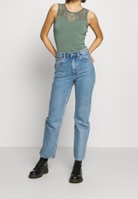 Grön ärmlös topp med spetsdetaljer, i kombination med högmidjade ljusblå jeans och svarta snörstövlar. Enkel, figursydd design.