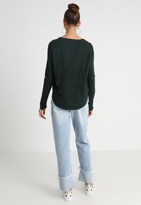 Mörkgrön stickad tröja med en avslappnad passform, matchad med ljusblå jeans med fransar och vita slip-on sneakers med svarta prickar.