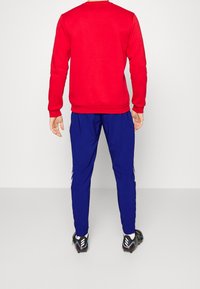 Czerwony sweter z długim rękawem, zestawiony z niebieskimi spodniami sportowymi z białymi paskami po bokach i czarnymi butami sportowymi.