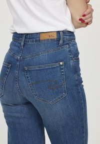 Gros plan sur une personne portant un jean bleu Lee Cooper avec des poches arrière visibles et un bras plié avec des ongles peints en rouge.