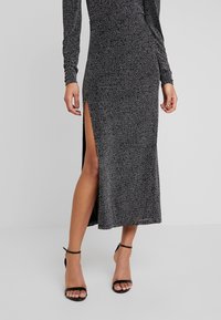 Žena v dlouhých černých šatech s bílými tečkami a rozparkem až ke stehnům, doplněná černými sandály na vysokém podpatku s páskem kolem kotníku.