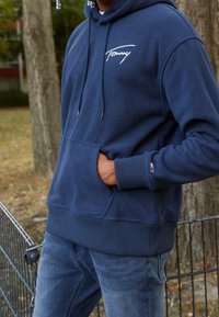 Marineblauwe fleece hoodie met een voorzak, capuchon met trekkoord en wit logo op de borst, voorzien van ribbelmanchetten en -zoom.