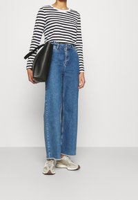 Schwarz-weiß gestreiftes Langarmhemd, hoch geschnittene, blaue, weite Denim-Jeans, graue und braune Sneakers und eine schwarze Schultertasche.