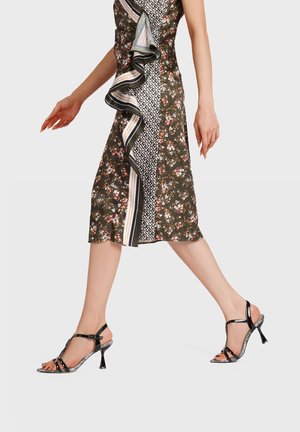 Donna in un vestito midi floreale e decorato che cammina, indossando sandali neri con tacco alto, cinturini alla caviglia e dita dei piedi scoperte.