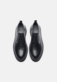Zapatos de cuero negro con cordones, con un acabado suave, punta redondeada y acentos de rayas horizontales en los costados. Suela de goma con bordes texturizados.