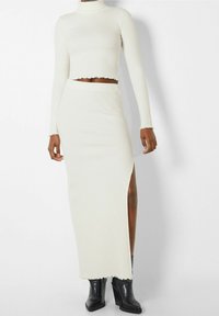 Femme portant un pull court en maille côtelée blanche à col roulé et une jupe taille haute assortie avec une fente latérale, accompagnée de bottines noires.