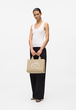 Γυναίκα με λευκό αμάνικο τοπ και μαύρο παντελόνι που κρατάει μπεζ τσάντα tote Karl Lagerfeld με λευκό κείμενο, στέκεται μπροστά σε λευκό φόντο.