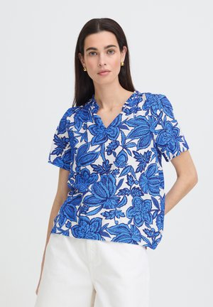 Femme portant un chemisier à manches courtes en V avec un grand motif floral bleu et un pantalon blanc, debout devant un fond clair uni.