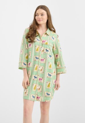 Mujer con un vestido camisé de color verde claro con mangas largas, que presenta un estampado colorido de animales y un bolsillo en el pecho.