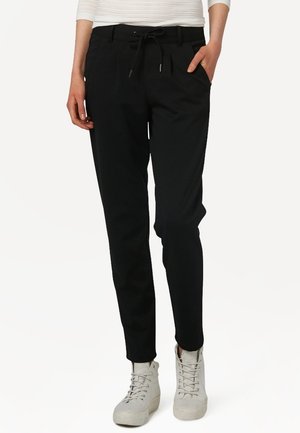 Personne portant un pantalon noir fuselé avec une taille à cordon et des baskets blanches montantes, debout avec une main dans la poche.