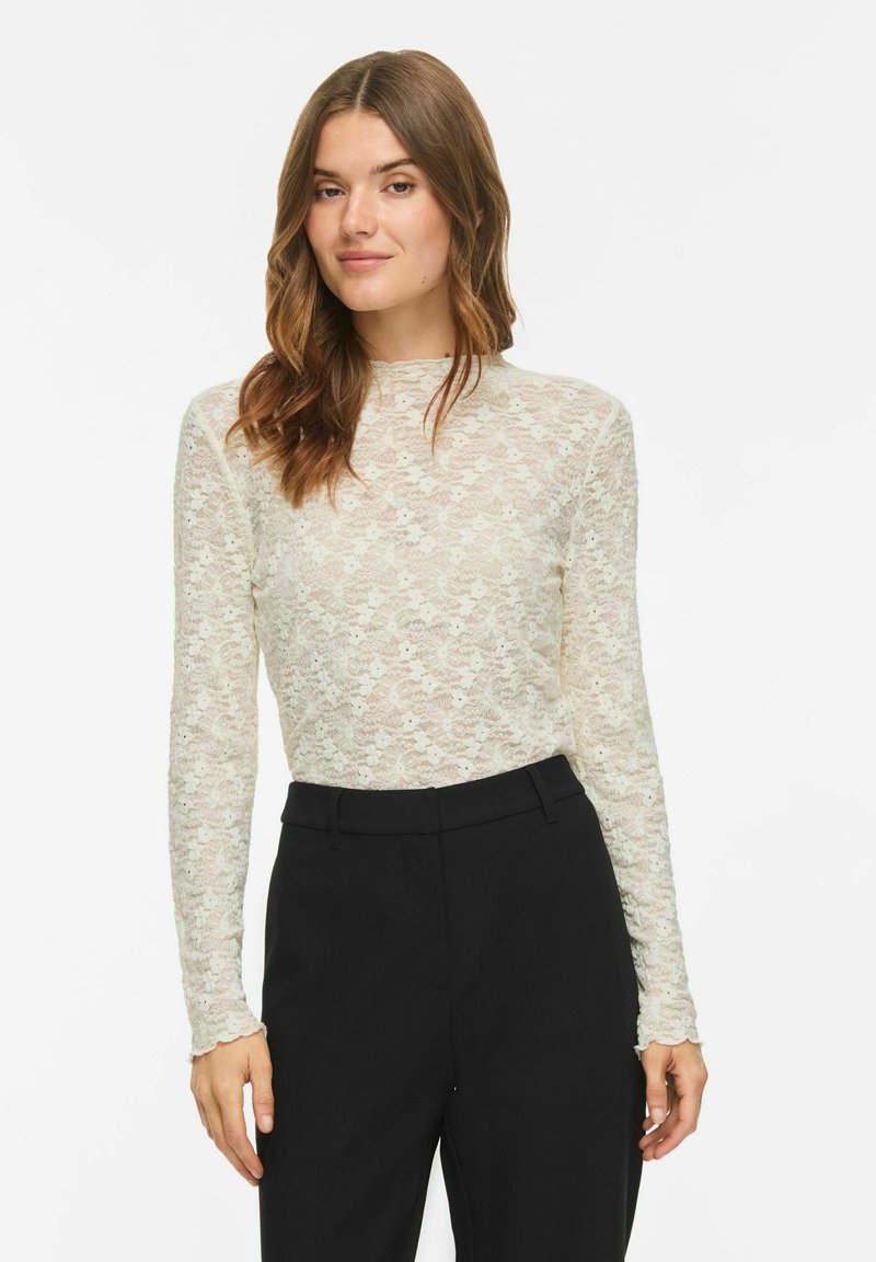 Femme aux longs cheveux bruns portant un top blanc en dentelle à manches longues et un pantalon noir taille haute, debout devant un fond uni.