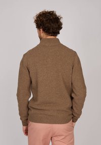 Pull en tricot marron avec un col montant, des poignets et un ourlet côtelés, mettant en valeur un motif de tissu texturé. Porté avec un pantalon rose pâle.