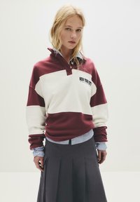 Giovane donna che indossa una maglietta rugby bordeaux e bianca con la scritta "New York City" sopra una camicia a righe, abbinata a una gonna plissettata nera, in piedi contro un muro bianco.
