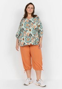 Flödande, mönstrad topp i turkos, orange, svart och vitt; kombinerad med orangea cropped byxor; inkluderar dragsko detaljer vid fållen.