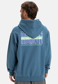 Sweat à capuche bleu avec une finition texturée. Présente une impression graphique en violet et vert au dos, mettant en valeur des motifs géométriques et des accents.