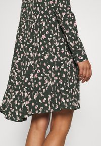 Blumenkleid in Grün mit verstreuten rosa und weißen Mustern. Lange Ärmel und ein plissiertes Design. Weicher Stoff und ausgestellter Saum.